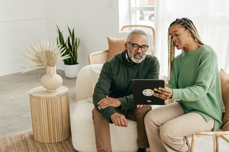 About Your Bill A woman sits with an elderly man on the couch showing him something on an iPad and explaining it to him