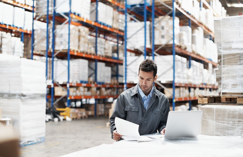 A male worker sits in a large warehouse with fully stocked shelves behind him as he compares notes while sitting at a laptop