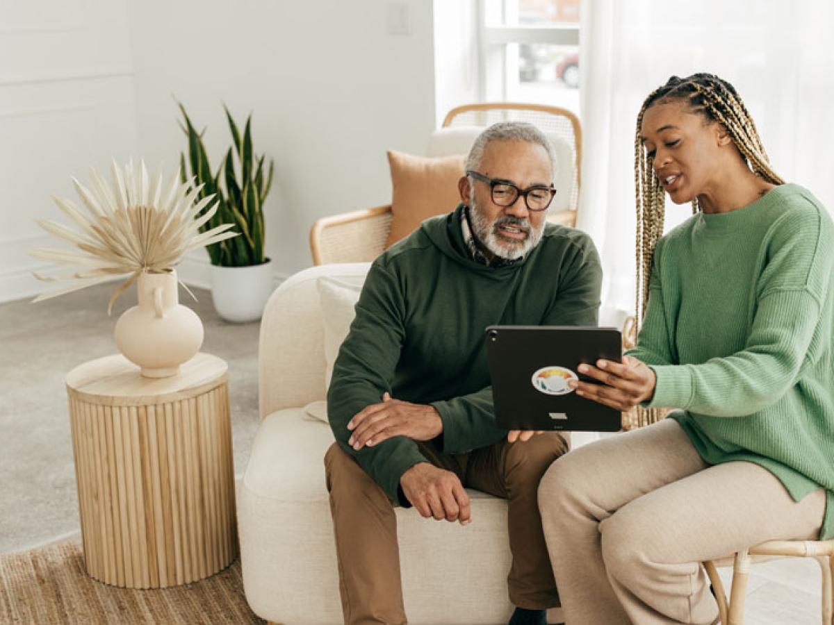 A woman sits with an elderly man on the couch showing him something on an iPad and explaining it to him