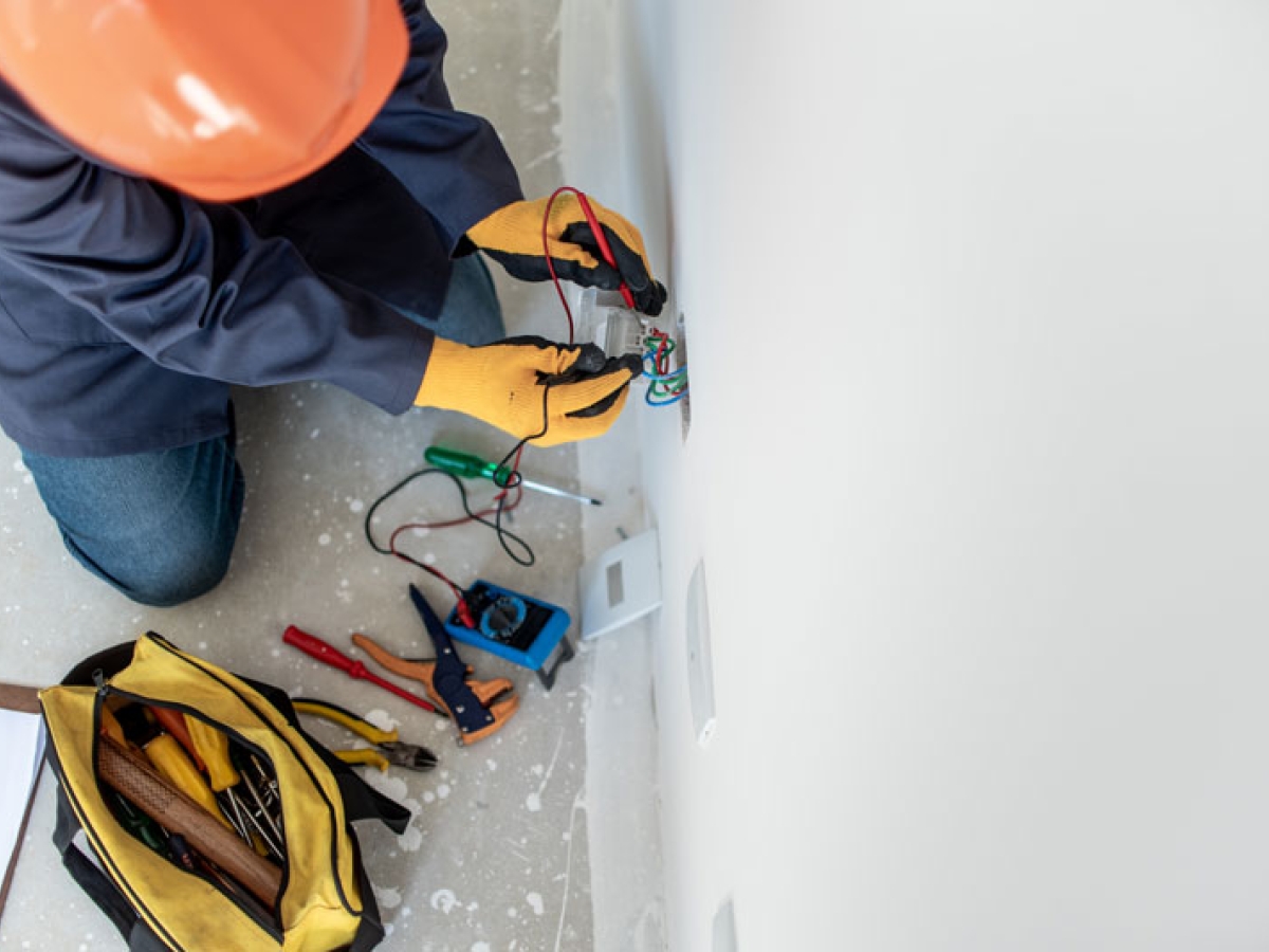 A worker in personal protective equipment has an open tool bag showing a number of tools is testing an outlet